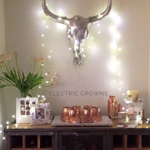 May include: A silver bull skull with horns hangs on a wall with white fairy lights strung around it. A wooden cabinet with a metal top has a set of copper mugs on a tray, two copper bottles, and two white candles. The text "ELECTRIC CROWNS" is printed on the wall.