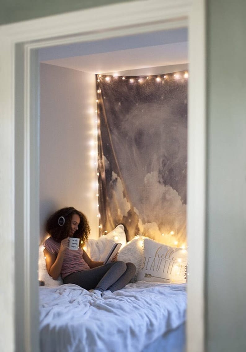 Op de afbeelding: Een gezellige slaaphoek met een wandtapijt van wolken en fee&euml;rieke lichtjes. Een persoon zit op een bed met kussens, leest een boek en houdt een mok vast. De kamer wordt omlijst door een deuropening.