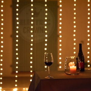 May include: A romantic table setting with two wine glasses, a bottle of wine, and a candle in a glass jar. The table is set on a white tablecloth and is illuminated by string lights hanging from the ceiling. The background features a sign that says "Electric Crowns".