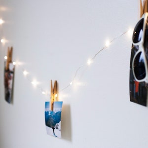 Puede incluir: Una pared blanca con una cadena de luces de hadas enhebradas a trav&eacute;s de ella. Las luces est&aacute;n sujetas a fotos y a un par de gafas de sol.