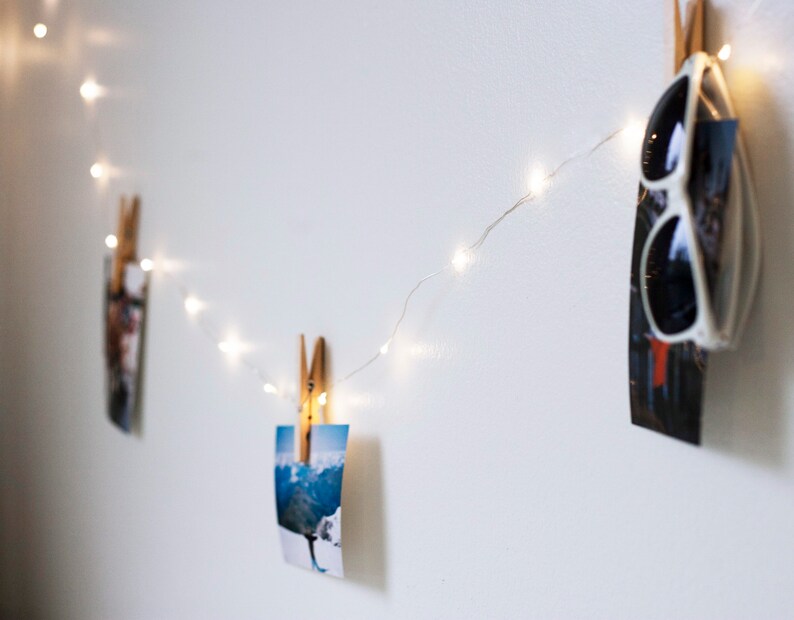 Puede incluir: Una pared blanca con una cadena de luces de hadas enhebradas a trav&eacute;s de ella. Las luces est&aacute;n sujetas a fotos y a un par de gafas de sol.