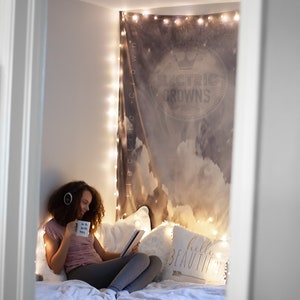 May include: A young person sits on a bed with a white blanket and pillows. They are wearing a pink shirt and grey pants. They are holding a mug that says "be the good vibes" and a tablet. The bed is decorated with string lights and a tapestry with the words "Electric Crowns" on it. There is a pillow with the words "hello beautiful" on it.