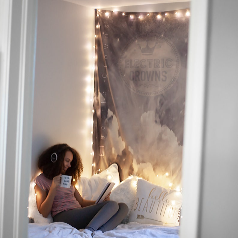 May include: A young person sits on a bed with a white blanket and pillows. They are wearing a pink shirt and grey pants. They are holding a mug that says "be the good vibes" and a tablet. The bed is decorated with string lights and a tapestry with the words "Electric Crowns" on it. There is a pillow with the words "hello beautiful" on it.