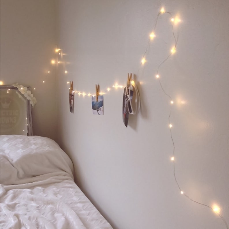 Puede incluir: Cama blanca con un edred&oacute;n blanco y una almohada blanca. Una cadena de luces de hadas est&aacute; colgada en la pared sobre la cama, con fotos sujetas a las luces con pinzas para la ropa.