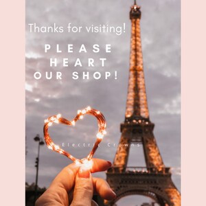 May include: A heart-shaped string of lights in front of the Eiffel Tower. The lights are lit and glowing. The text "Electric Crowns" is visible below the heart.