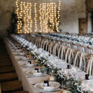 May include: A long table set for a wedding reception with white linens, wooden chairs, and a string of lights hanging above. The table is decorated with greenery and candles.
