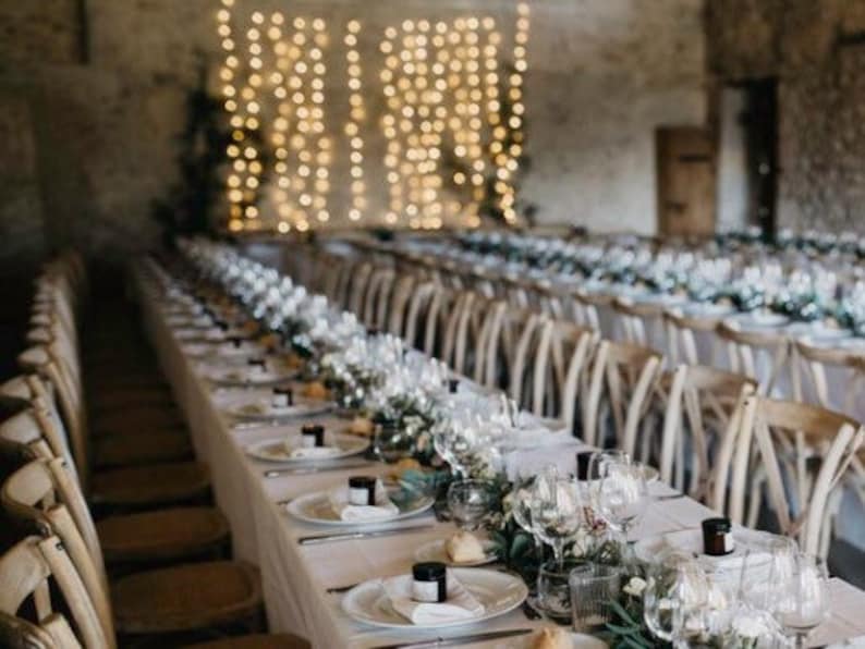 May include: A long table set for a wedding reception with white linens, wooden chairs, and a string of lights hanging above. The table is decorated with greenery and candles.