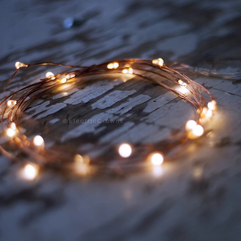 May include: Copper wire fairy lights with warm white LED bulbs, arranged in a circle on a white wooden surface. The lights are strung together with a thin copper wire, and the bulbs are spaced evenly apart. The lights are turned on, and the bulbs are glowing brightly. The image includes the text "@ElectricCrowns".