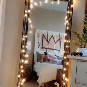 Peut inclure: Un miroir de pleine longueur avec un cadre noir et une guirlande de lumi&egrave;res blanches scintillantes enroul&eacute;es autour du cadre. Le miroir refl&egrave;te une chambre &agrave; coucher avec un lit, une personne allong&eacute;e sur le lit et une &oelig;uvre d'art au mur.