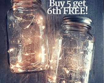 May include: Two clear glass mason jars with silver lids filled with white fairy lights. The jars are on a rustic wooden background. The text "Buy 5 get 6th FREE!" is written in white on a black background.