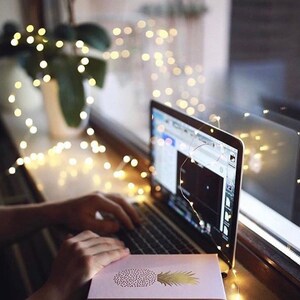 Op de afbeelding: Een laptop met een scherm waarop een webpagina wordt weergegeven, met een roze kaart met een gouden ananasontwerp. Slingers zijn rond de laptop en het raam gedrapeerd. Een plant in een witte vaas is zichtbaar op de achtergrond.