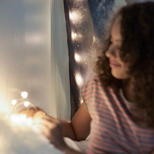 Puede incluir: Una persona joven con cabello casta&ntilde;o rizado se sienta en una cama con luces de cadena. La persona lleva una camisa a rayas y sostiene las luces de cadena en sus manos.