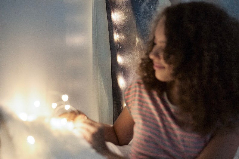 Puede incluir: Una persona joven con cabello casta&ntilde;o rizado se sienta en una cama con luces de cadena. La persona lleva una camisa a rayas y sostiene las luces de cadena en sus manos.