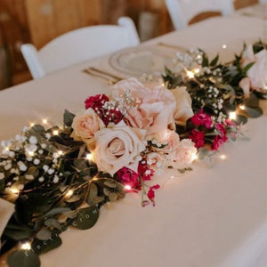 May include: A wedding table centrepiece with a garland of greenery, blush pink roses, and burgundy carnations. The garland is accented with a string of fairy lights.