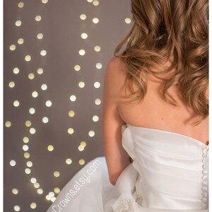 May include: A bride wearing a white strapless wedding dress with a beaded embellishment on the skirt. The dress has a fitted bodice and a flowing skirt. The bride is standing in front of a wall with string lights.