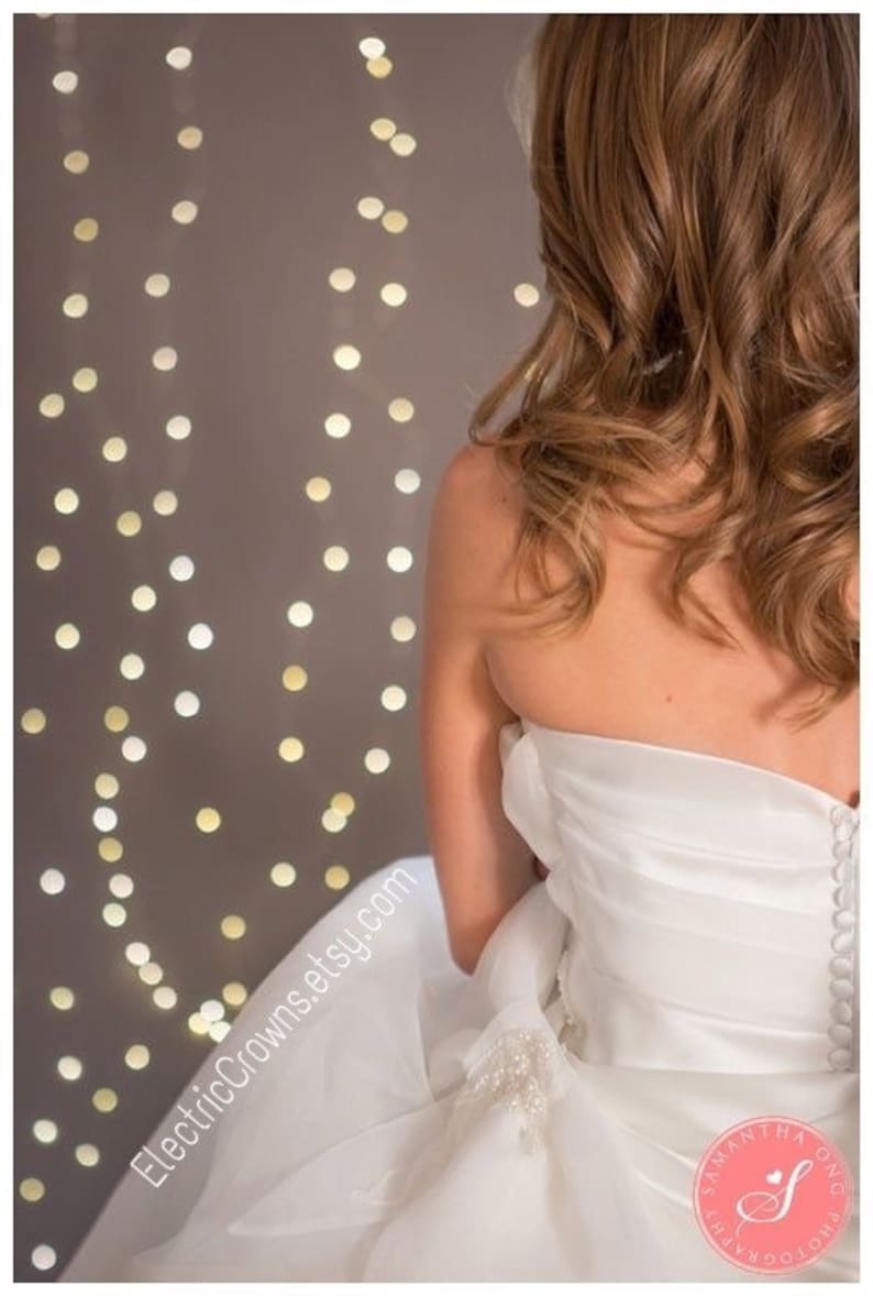 May include: A bride wearing a white strapless wedding dress with a beaded embellishment on the skirt. The dress has a fitted bodice and a flowing skirt. The bride is standing in front of a wall with string lights.