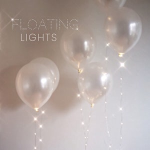 Könnte beinhalten: Weiße Ballons mit Lichterketten im Inneren, die vor einem weißen Hintergrund schweben. Der Text "FLOATING LIGHTS" befindet sich oben im Bild.