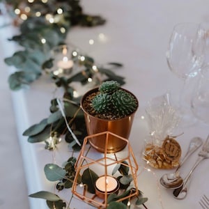 May include: A copper geometric candle holder with a small white candle inside. The candle holder is surrounded by greenery and fairy lights. A small copper pot with a succulent plant is in the background.