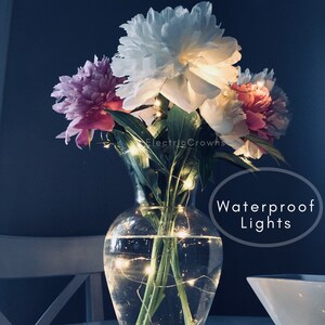 May include: A clear glass vase filled with water and white and pink peonies.  A string of warm white fairy lights is wrapped around the stems of the flowers. The text "Waterproof Lights" is written in a white circle on the image.