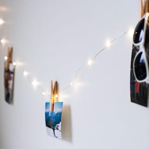 May include: A white wall with a string of fairy lights strung across it.  The lights are clipped to photos and a pair of sunglasses.