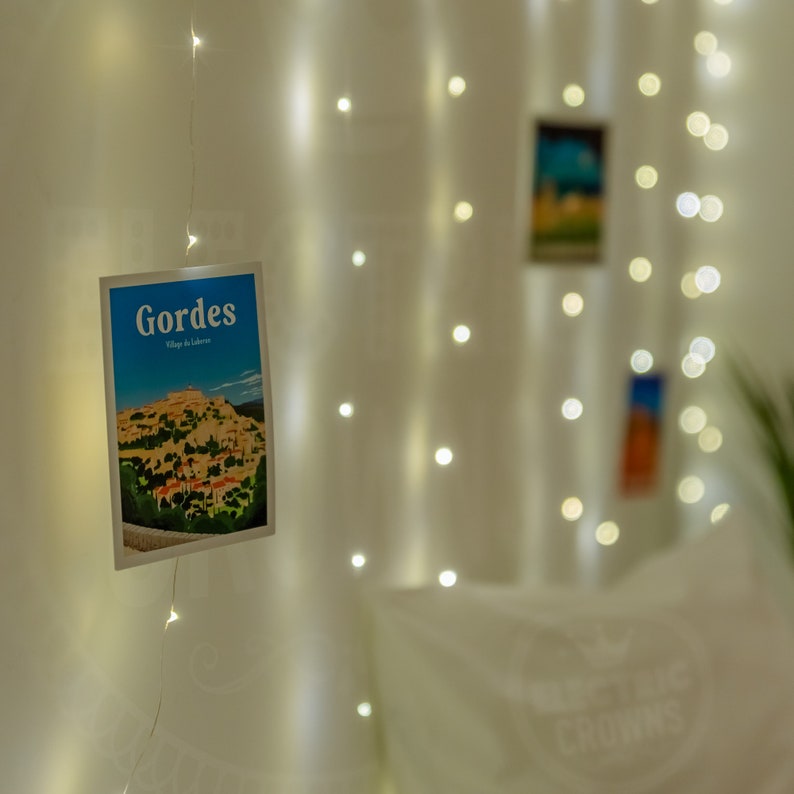 May include: A blue postcard with the text "Gordes Village de la Luberon" and a picture of a village with buildings and trees. The postcard is hanging on a white wall with white fairy lights.