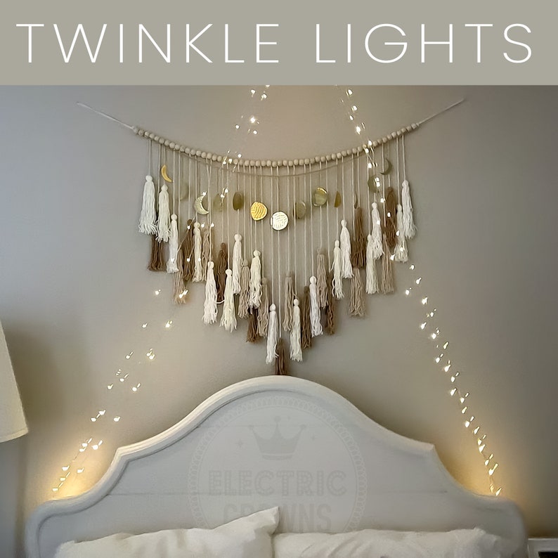 May include: A white headboard with a gold crown logo and the text "Electric Dreams" on it. A white and beige macrame wall hanging with gold moon shapes is above the headboard. A string of fairy lights is draped over the headboard and wall hanging.
