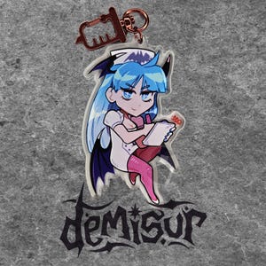 May include: A blue and pink anime-style character with bat wings and a nurse's outfit holding a clipboard. The character is wearing a white nurse's hat with a bat on top. The character is on a clear acrylic keychain with the word "DEMISUR" printed below.