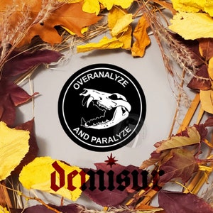 May include: A round black sticker with white text that reads "OVERANALYZE AND PARALYZE" surrounds a white skull graphic. The sticker is surrounded by autumn leaves in shades of yellow, orange, and red.