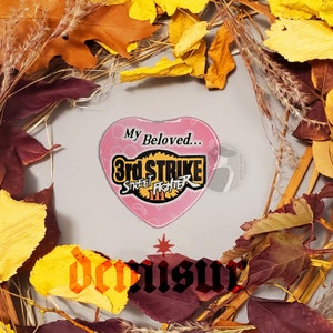 May include: A pink heart-shaped pin with the text "My Beloved... 3rd Strike Street Fighter III" in yellow and black lettering.