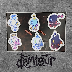 May include: A sheet of stickers featuring six cartoon characters with angel and devil themes. The stickers are in various colors, including blue, pink, and black, with the word "demisur" printed on the sheet.