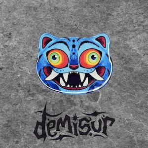 May include: A blue cat-shaped graphic with large, colorful eyes and sharp teeth. The design features red accents and a white outline. Below the graphic is the word "Demisur" in a stylized font, set against a textured gray background.
