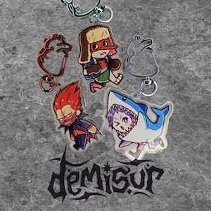 May include: A collection of acrylic keychains featuring cartoon characters and heart-shaped designs. The keychains include a boxer, a shark, and a character with red hair. The word "demisur" is printed in a stylized font.