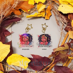 May include: Two acrylic keychains featuring a cartoon character with the text "Skill Issue." One keychain is holographic, the other is normal. Both have gold star-shaped attachments. The keychains are surrounded by autumn leaves.