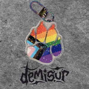 May include: A clear acrylic paw print keychain with a rainbow and pride flag design. The keychain has a silver-colored clasp and a decorative star pattern. The text "Demisur" is at the bottom of the image.