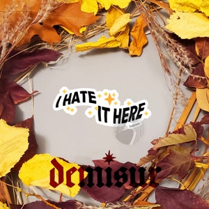 May include: A sticker with the text "I hate it here" in black text on a white background with yellow stars. The sticker is placed on a background of fall leaves in shades of brown, orange, and red.