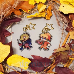 May include: Two character charms featuring a green and yellow lizard holding a knife and a blonde character holding a plate of food. The charms are attached to gold star-shaped keychains.