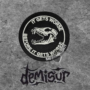 May include: Black circular patch with white text that reads "IT GETS WORSE BEFORE IT GETS WORSE" around a white skull graphic. Below the patch is a black gothic-style logo that reads "Demisur".