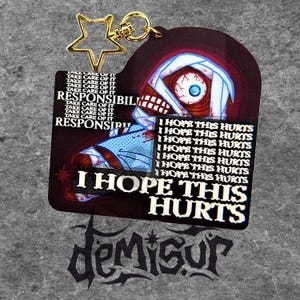 May include: A dark red and black keychain with a graphic of a bandaged skull with a single eye. The keychain has the text "I HOPE THIS HURTS" and "RESPONSIBILITY". A gold star-shaped charm and a gold clasp are attached.