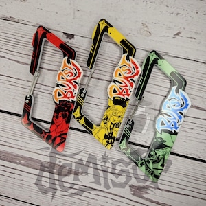 May include: Three colorful carabiners with anime-style character art. The carabiners are red, yellow, and green, each with a different design. The designs include stylized text and character illustrations. The carabiners are made of metal and plastic.