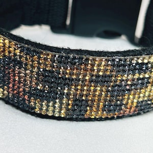 May include: A black collar featuring a dense, multi-colored pattern of small, reflective crystals in shades of gold, amber, and black. The collar includes a black fabric backing and a black plastic side-release buckle for secure fastening.