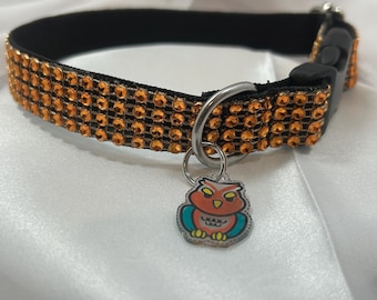 Puede incluir: Un collar para mascota de color negro con cuatro filas de pedrería naranja, una hebilla de plástico negro, una anilla metálica plateada y un pequeño dije de acrílico con forma de búho y detalles en naranja, turquesa y amarillo.