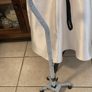 Könnte beinhalten: Ein silberner Gehstock mit schwarzem Griff und einem vierbeinigen Standfuß. Der Stock ist mit Strasssteinen besetzt.