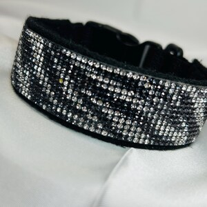 May include: A black collar featuring a wide band covered in numerous small, reflective silver-toned rhinestones arranged in a dense grid pattern. The collar includes a black plastic side-release buckle closure for securing the ends together.