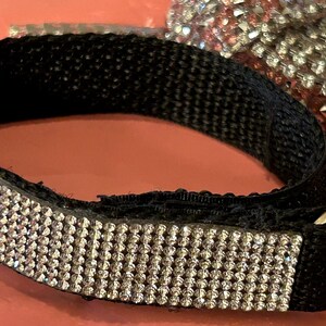 May include: Black wrist strap with a silver metal ring and a band of sparkling rhinestones. The strap is made of black fabric and has a decorative rhinestone band. A second, similar item is visible in the background.
