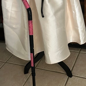 May include: A black and pink walking cane with a padded handle and a wrist strap. The cane is decorated with pink rhinestones.