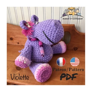 Può includere: Un ippopotamo di peluche viola lavorato a uncinetto con una sciarpa rosa. Il giocattolo è seduto su una superficie di legno. Il testo "Violette" è visibile sotto il giocattolo. Il testo "Patron/Pattern PDF" è visibile sotto il giocattolo. Il testo "Vivi Tricote! Oursons et Compagnons" è visibile nell'angolo in alto a destra dell'immagine.
