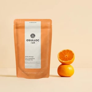 Może przedstawiać: Pomarańczowe opakowanie herbaty z napisem "OSULLOC" i "JEJU TANGERINE BLENDED TEA". Połówka pomarańczy leży na całej pomarańczy. Tło jest w jasnobeżowym kolorze.