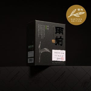 May include: A black box of tea with Korean text and a leaf illustration. The box has a white label with additional text and a gold circular emblem that reads "HAETCHA EDITION". The box is sitting on a black surface.