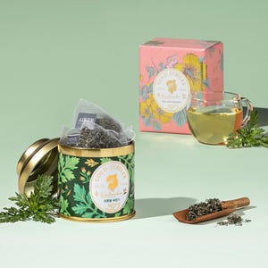 Può includere: Un set da tè con una scatola verde e oro con bustine di tè, una scatola floreale rosa, una tazza di vetro con tè e un cucchiaio di legno con tè sfuso. La scatola e il cofanetto riportano le parole "Gold Jubilee" e un logo. Il tè è di colore verde chiaro.