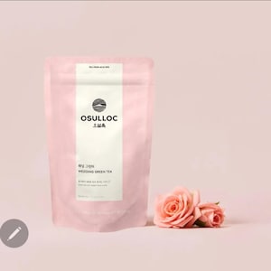 May include: A pink tea package with the brand name "Osulloc" and the text "Wedding Green Tea." Two pink roses are next to the package. The background is a soft pink.